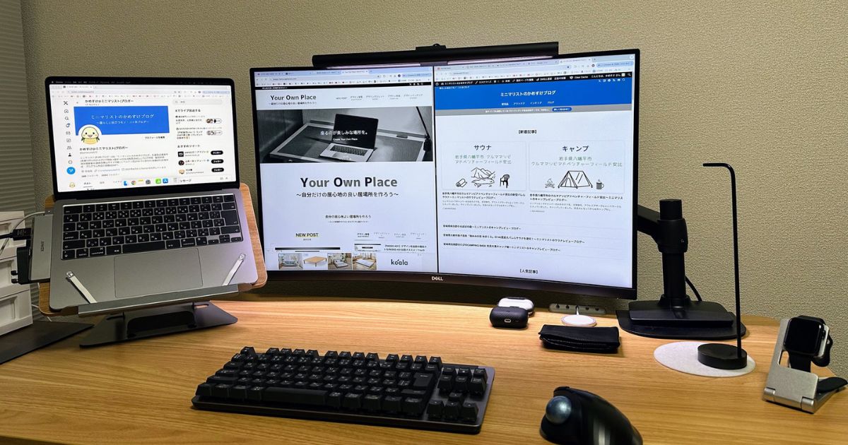 木製の机の上に置かれたデュアルモニターのワークスペース。モニターにはウェブサイトが表示されており、ノートパソコンがスタンドに設置されている。キーボード、マウス、その他の周辺機器も整頓されている。
